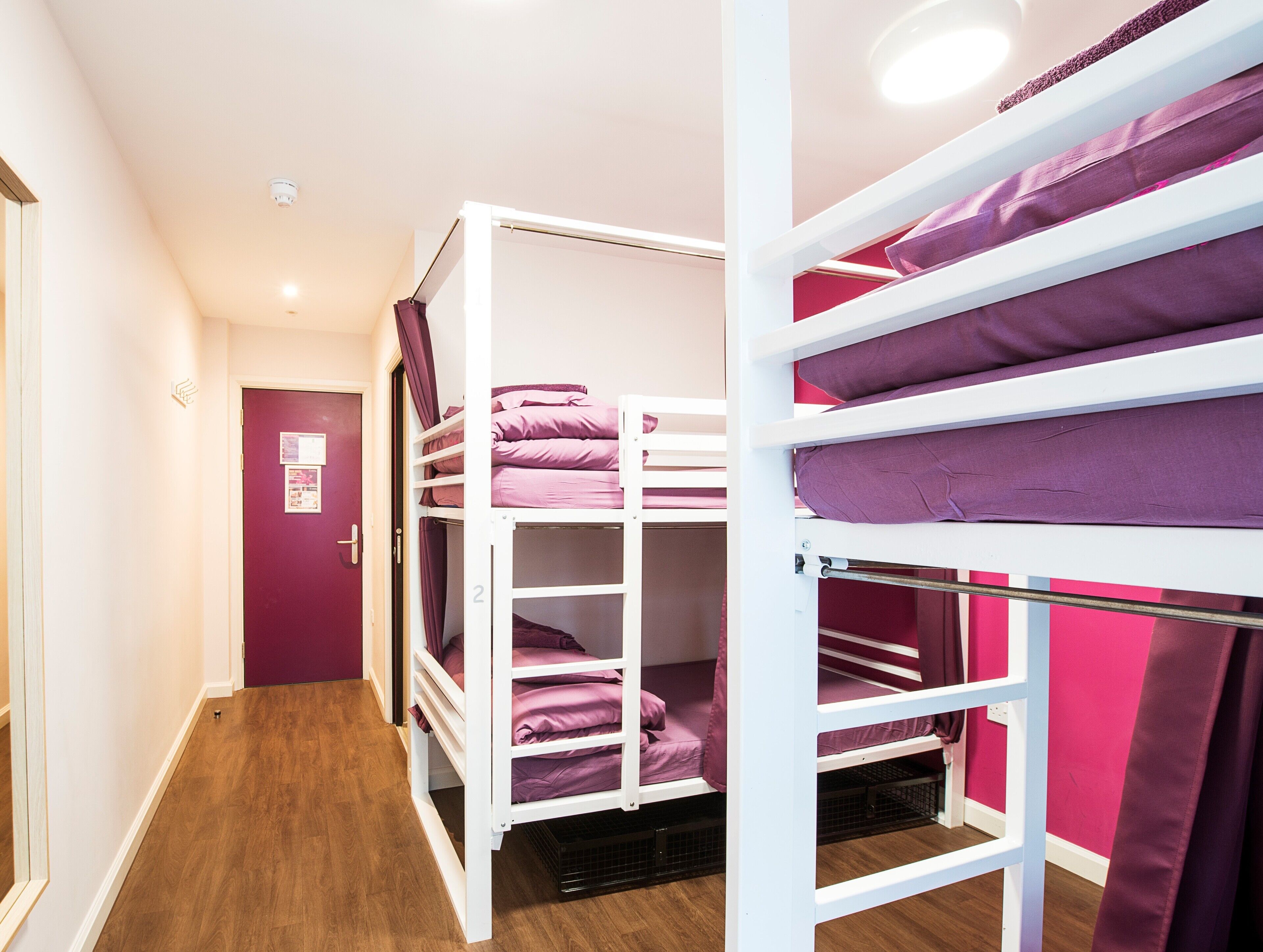 A dormitory room with bunk beds, wooden flooring, and a door with a bulletin board.