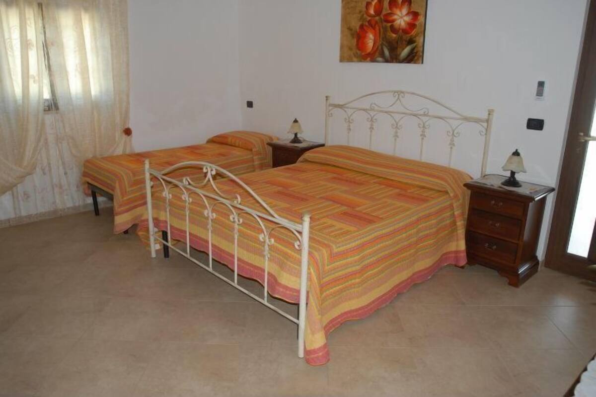 Une chambre avec deux lits, une fenêtre avec des rideaux, un tableau au mur et une table de chevet en bois avec une lampe.