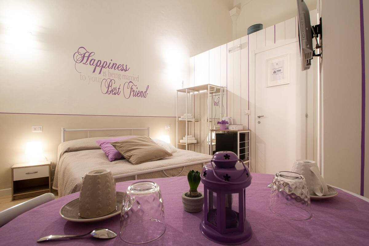 Ein Schlafzimmer mit einem Bett, Nachttischen, einem violetten Tisch mit Dekoration und einer Garderobe. An der Wand ist ein Spruch zu lesen: 'Happiness is being married to your Best Friend'.
