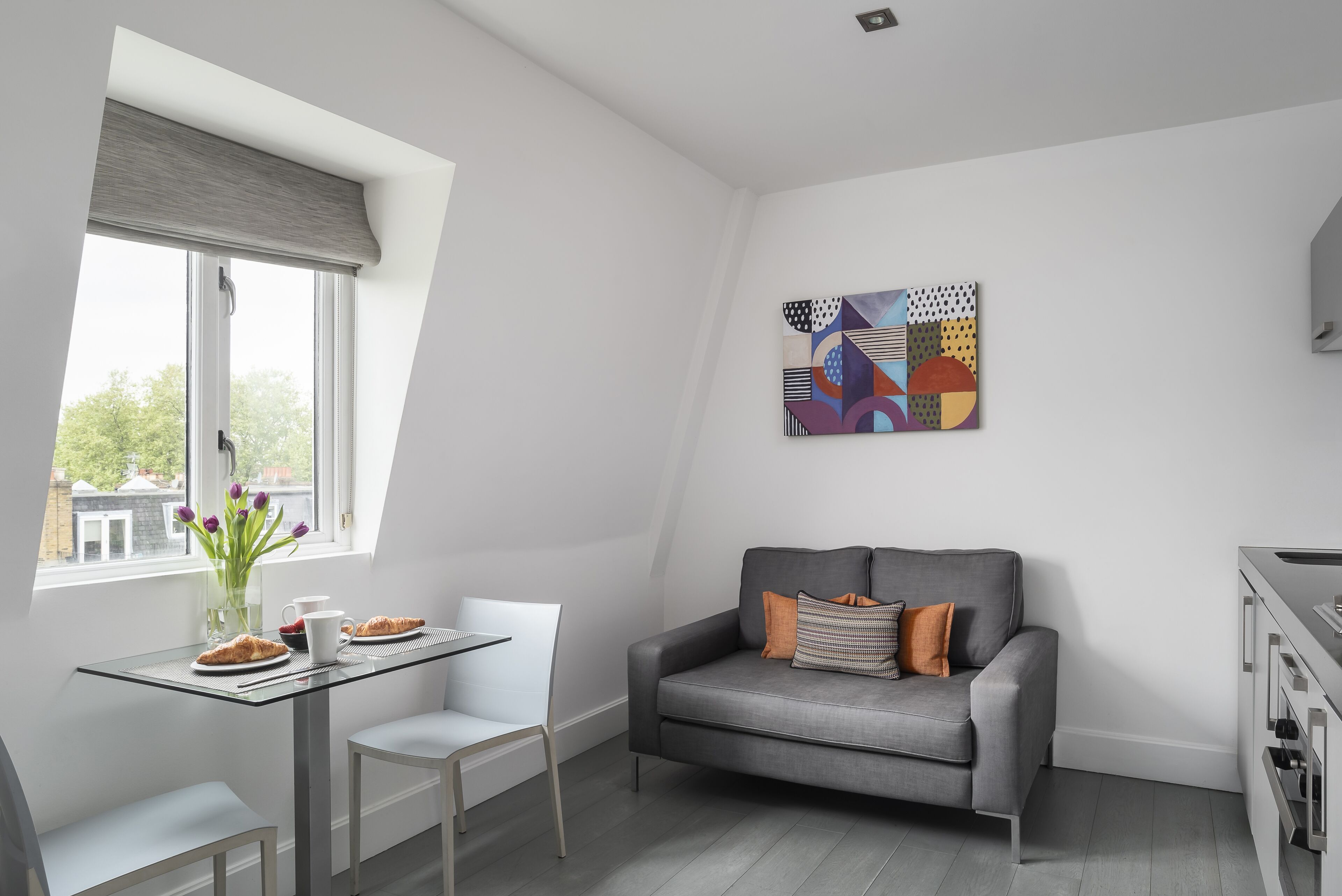 A modern living room with a grey sofa, a glass table with croissants, a white chair, and a painting on the wall.