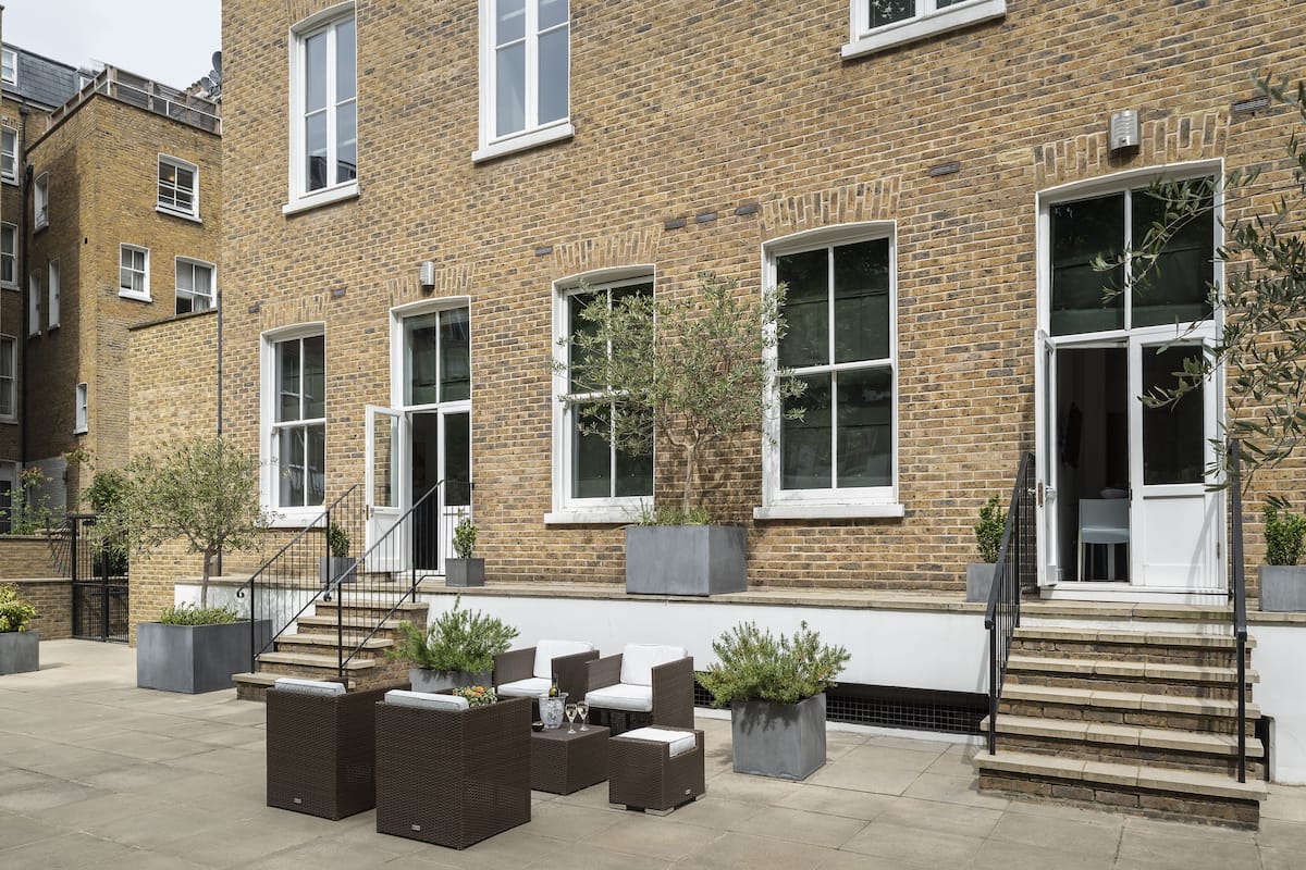 A brick building with a staircase leading to a door, outdoor seating area with wicker furniture, and potted plants.