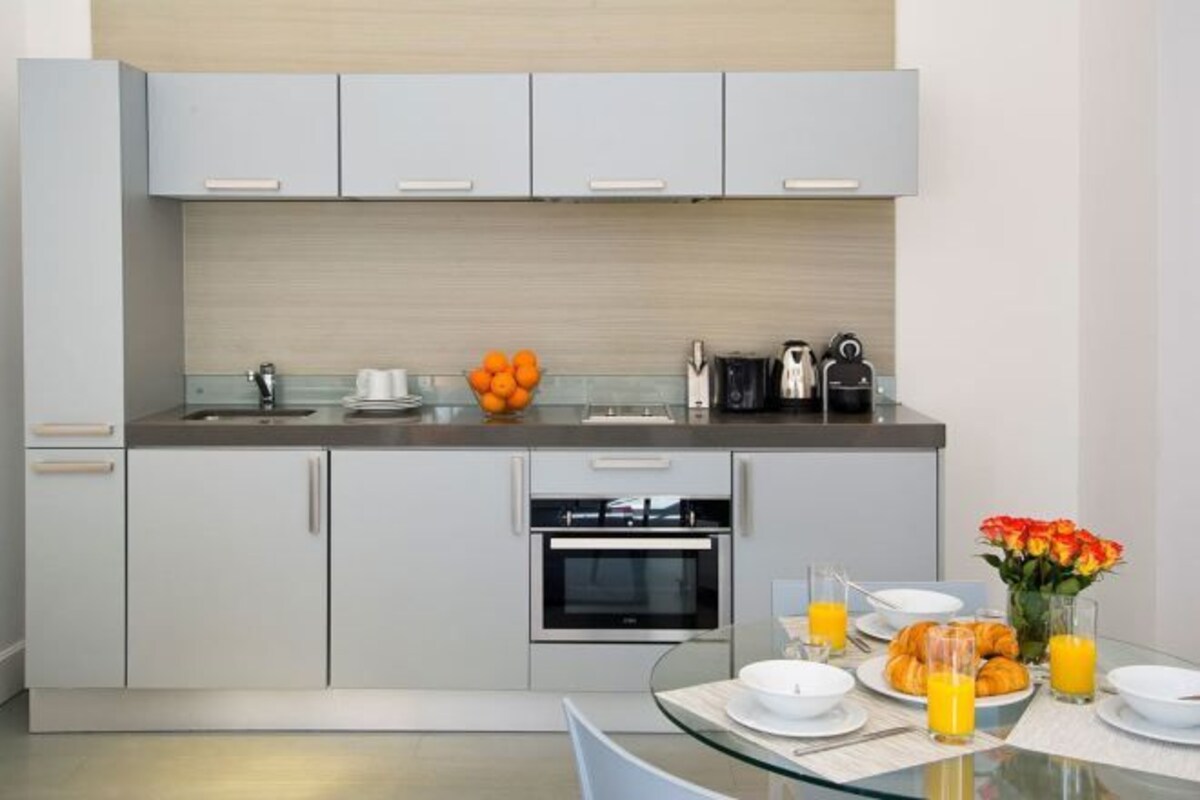 A modern kitchen with grey cabinets, a built-in oven, and a glass dining table set with plates, glasses, and a fruit bowl.