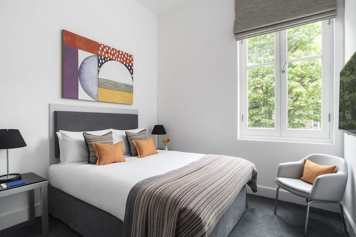 A neatly arranged hotel room with a large bed, two bedside tables with lamps, a chair, a window with a view of trees, and a piece of abstract art on the wall.