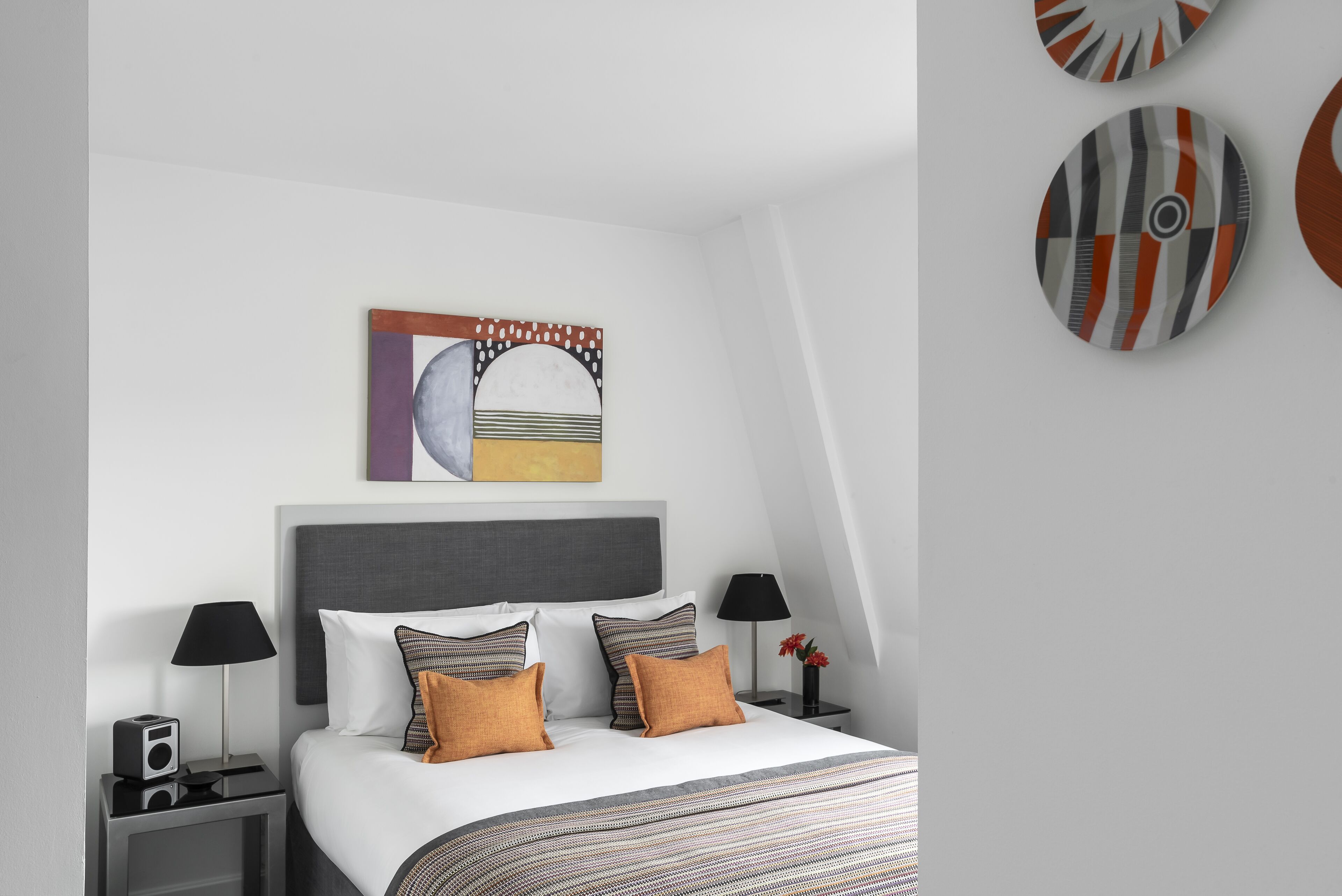 A modern hotel room with a neatly made bed, two bedside tables with lamps, a wall-mounted artwork, and a circular mirror on the wall.