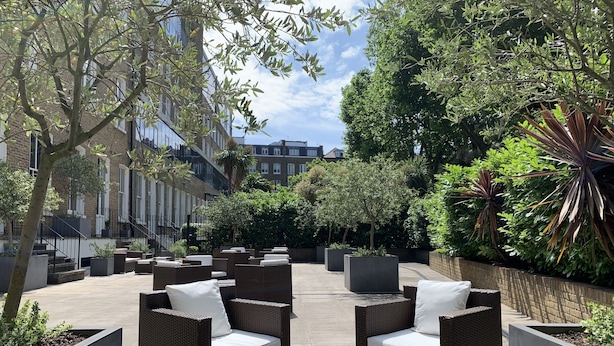An outdoor seating area with wicker furniture and planters, surrounded by trees and buildings. - Templeton Place by Supercity Aparthotels (London)