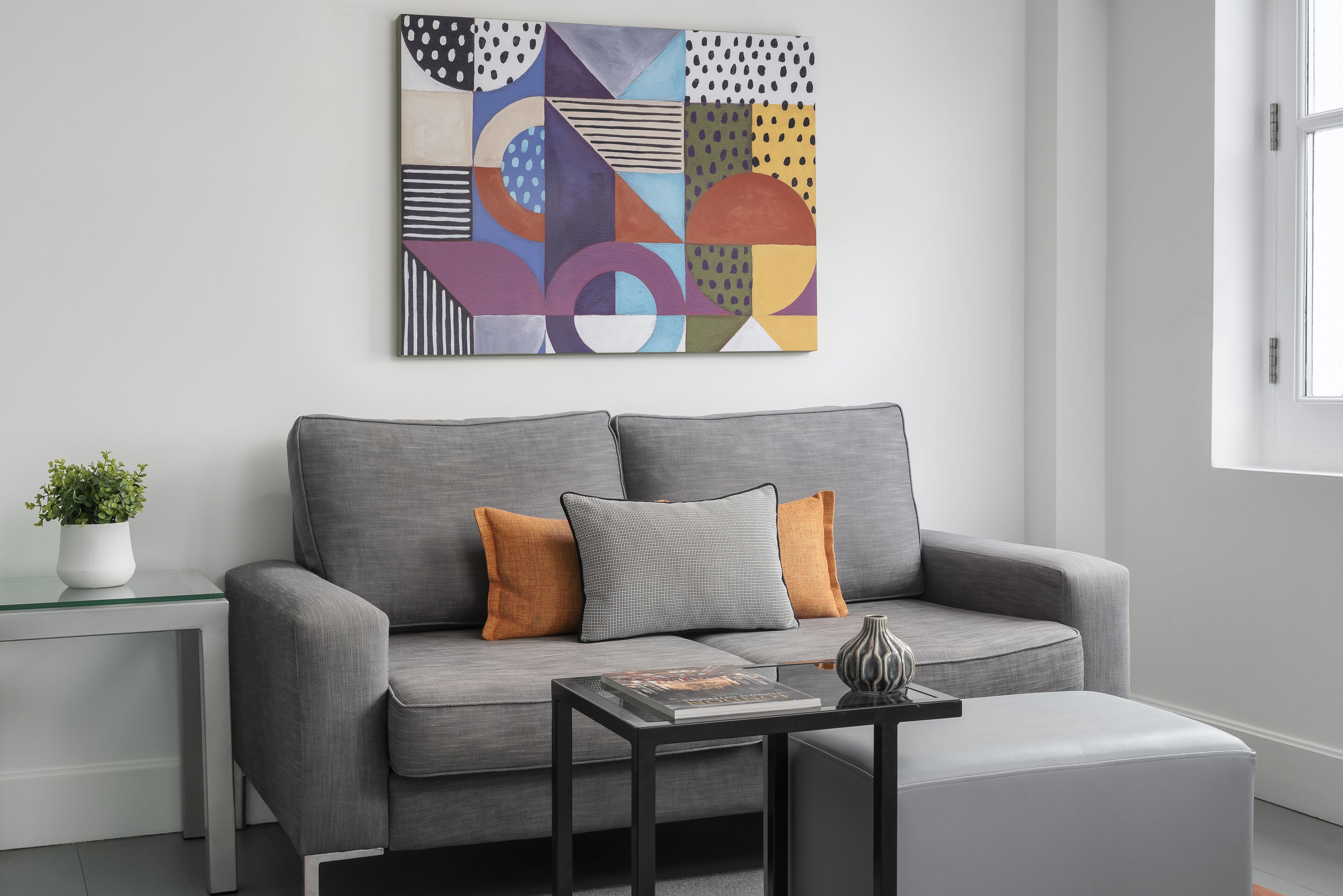 Una sala de estar moderna con un sofá gris, dos cojines, una mesa de centro de vidrio y un cuadro abstracto geométrico en la pared.