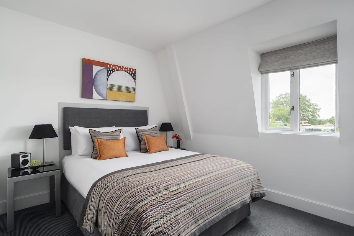 A modern hotel room with a large bed, a nightstand with a radio, a painting on the wall, and a window with a view of trees.