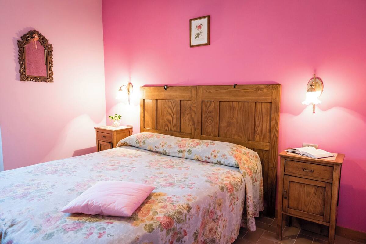Une chambre à coucher avec une tête de lit en bois, une literie à motifs floraux, un coussin rose, deux tables de chevet avec des lampes et un tableau encadré au mur.