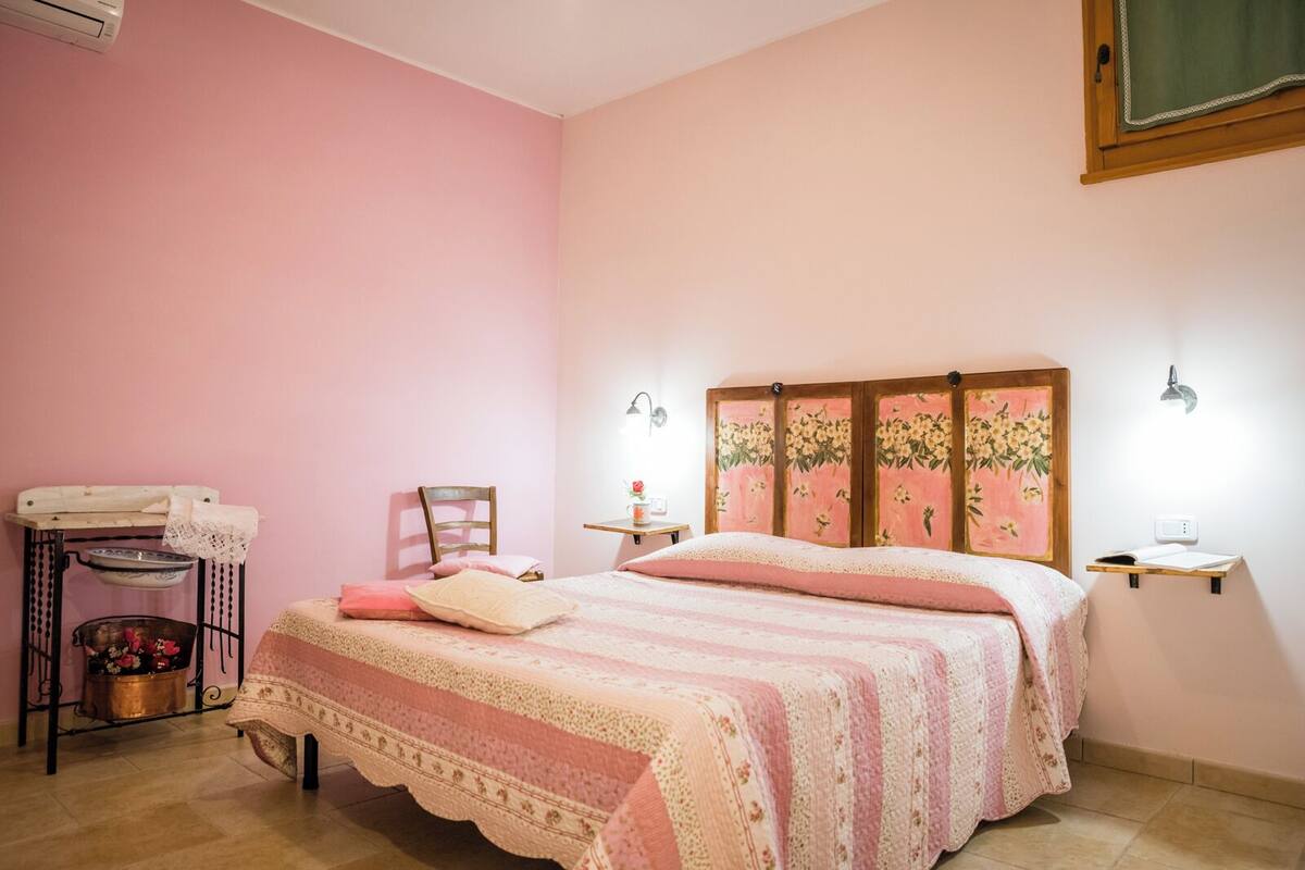 Une chambre avec un mur rose, un lit doté d’une tête de lit à motifs floraux, une chaise, une petite table avec une lampe et une étagère avec un livre.