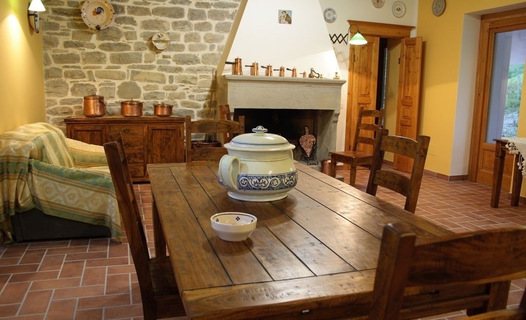 A rustic dining room with a wooden table, chairs, a large ceramic pot, and a fireplace.