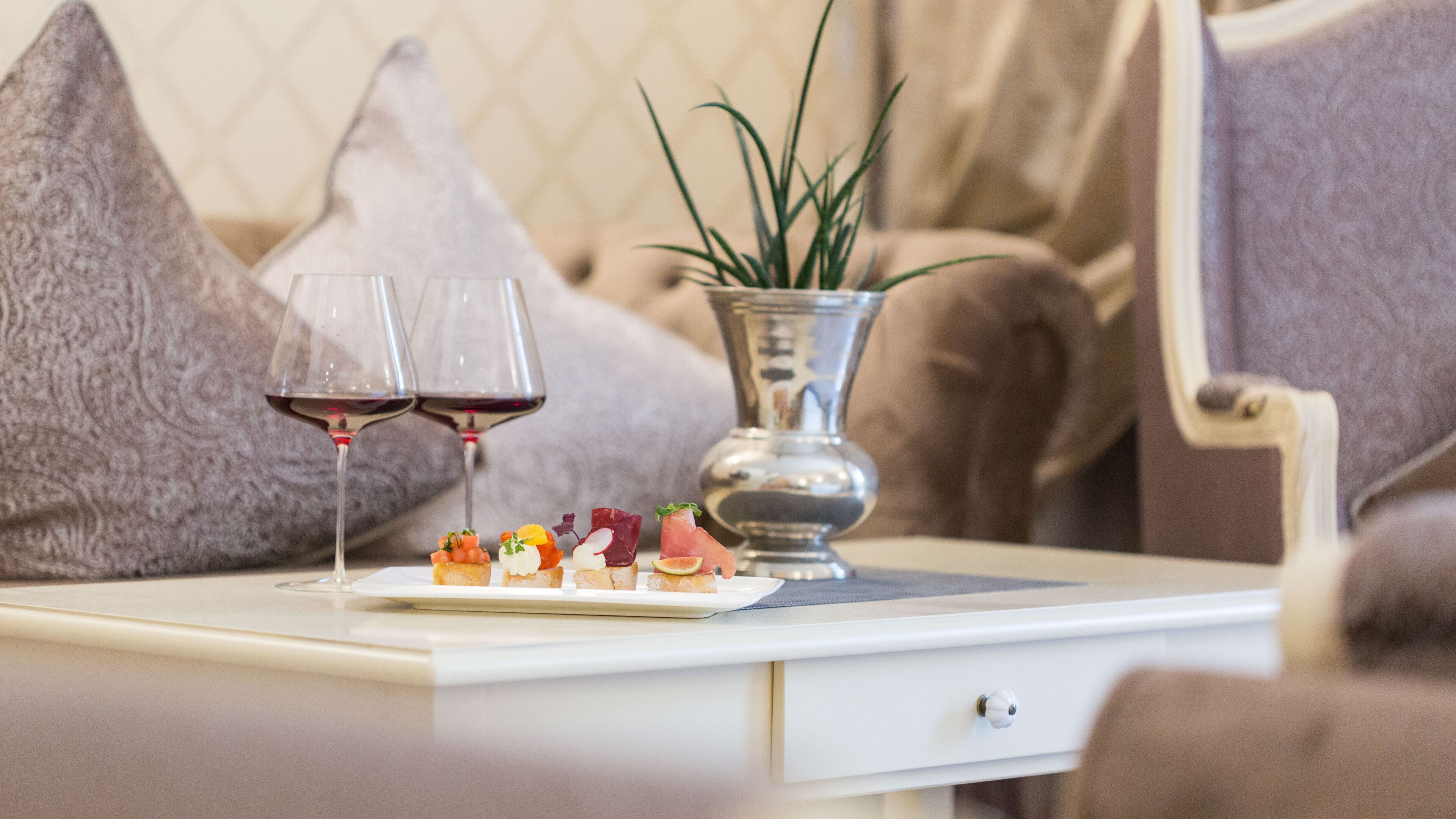A dining table with a white tablecloth, two glasses of red wine, and a plate of appetizers.