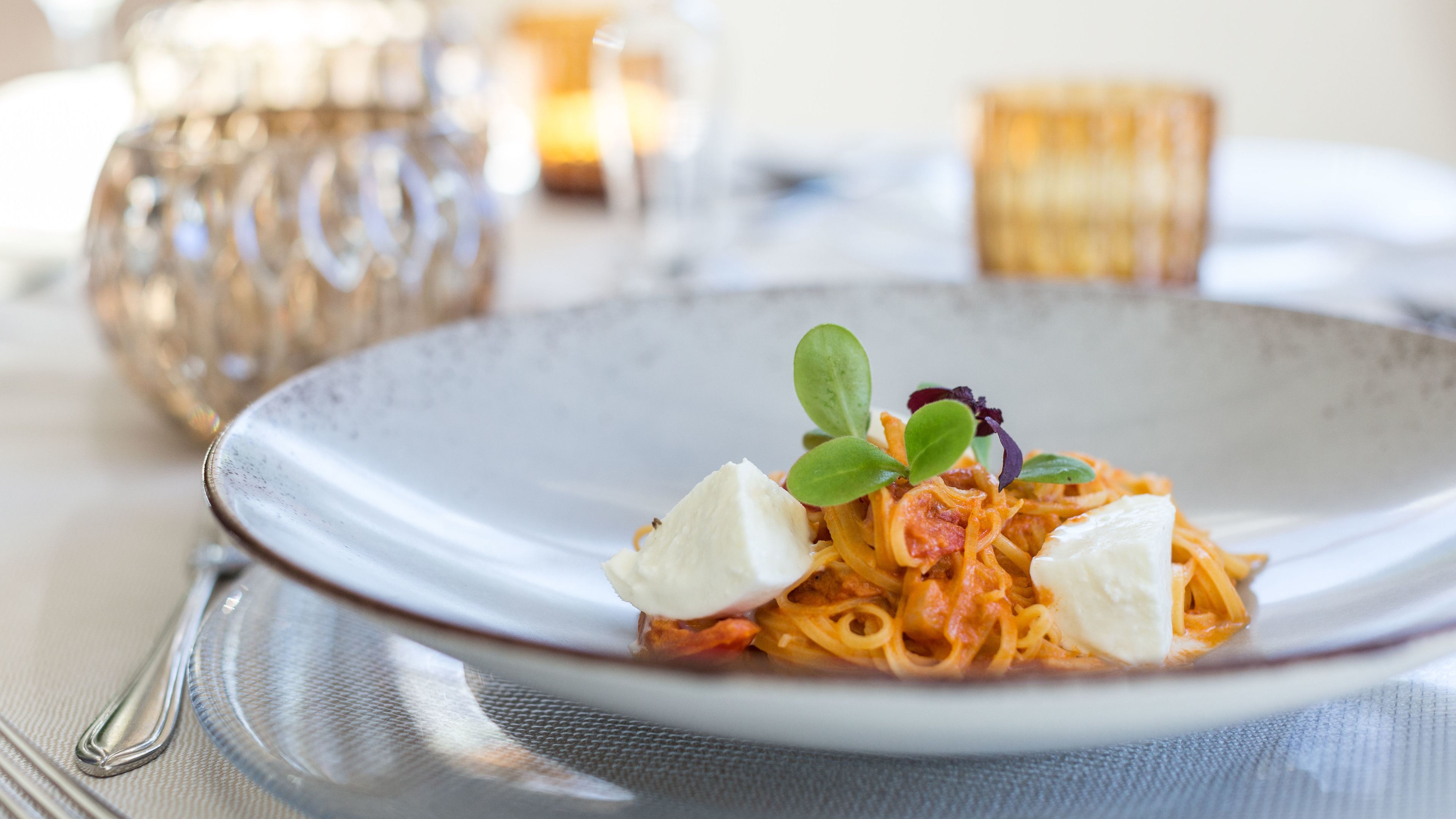 Une assiette de pâtes agrémentée d’une garniture, un récipient en verre et une bougie en arrière-plan.