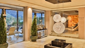 A modern hotel lobby with a glass wall offering a view of outdoor seating and greenery. There are potted plants, a seating area with a table, and a unique spherical chandelier. - Hotel Rössl (Parcines)