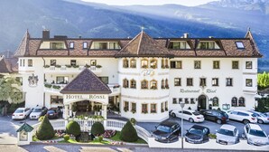 A hotel with a traditional European architectural style, featuring a prominent entrance, multiple balconies with flower boxes, and a parking area with several cars. - Hotel Rössl (Parcines)