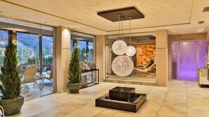 A modern hotel lobby with a fireplace, seating area, and large windows offering a view of the outdoors. - Hotel Rössl (Parcines)