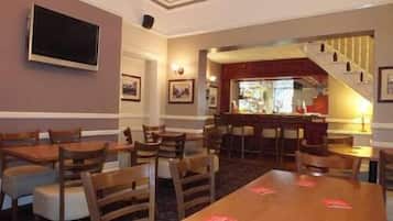 A hotel dining area with multiple wooden tables and chairs, a bar area with stools, and framed pictures on the walls.
