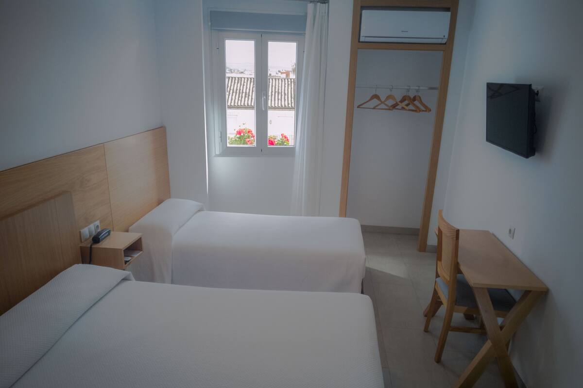 A hotel room with two beds, a desk, a chair, a TV, a wardrobe, and a window with a view of buildings.