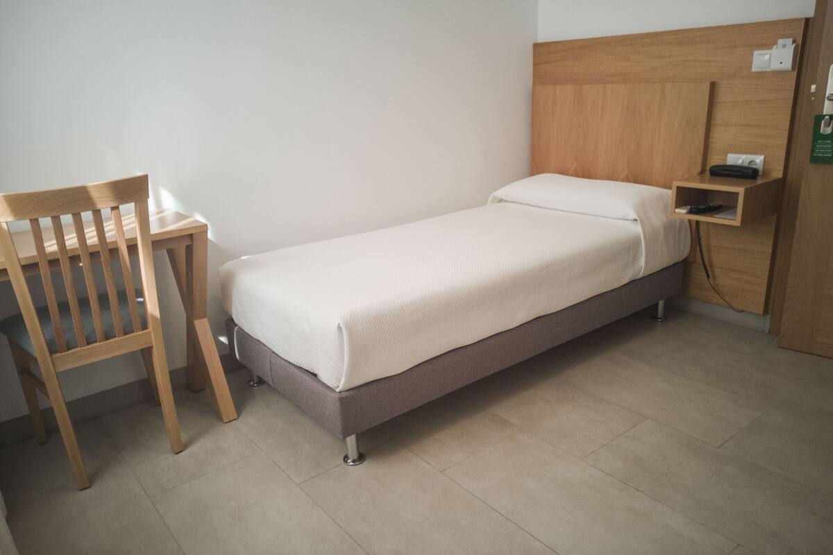 A single bed with a wooden headboard, a bedside table, and a chair in a hotel room.