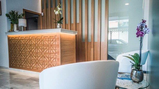 A reception area with a wooden reception desk, a white chair, and a glass table with a plant. - Abadi (Córdoba)
