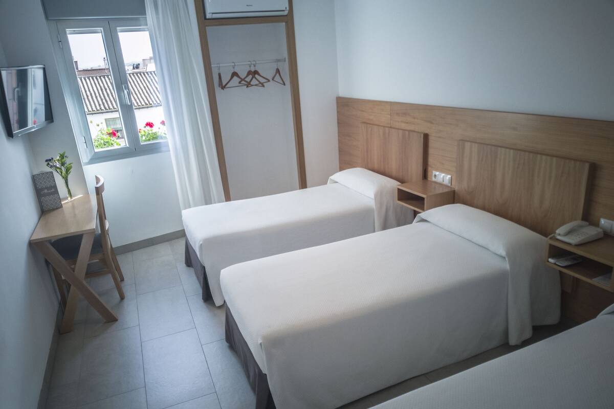 A hotel room with two single beds, a small desk with a chair, a TV mounted on the wall, and a window with a view of the outside.