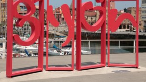 Una escultura roja con la palabra "Bergin" en una fuente estilizada, ubicada en un entorno urbano con edificios y un frente marítimo al fondo.