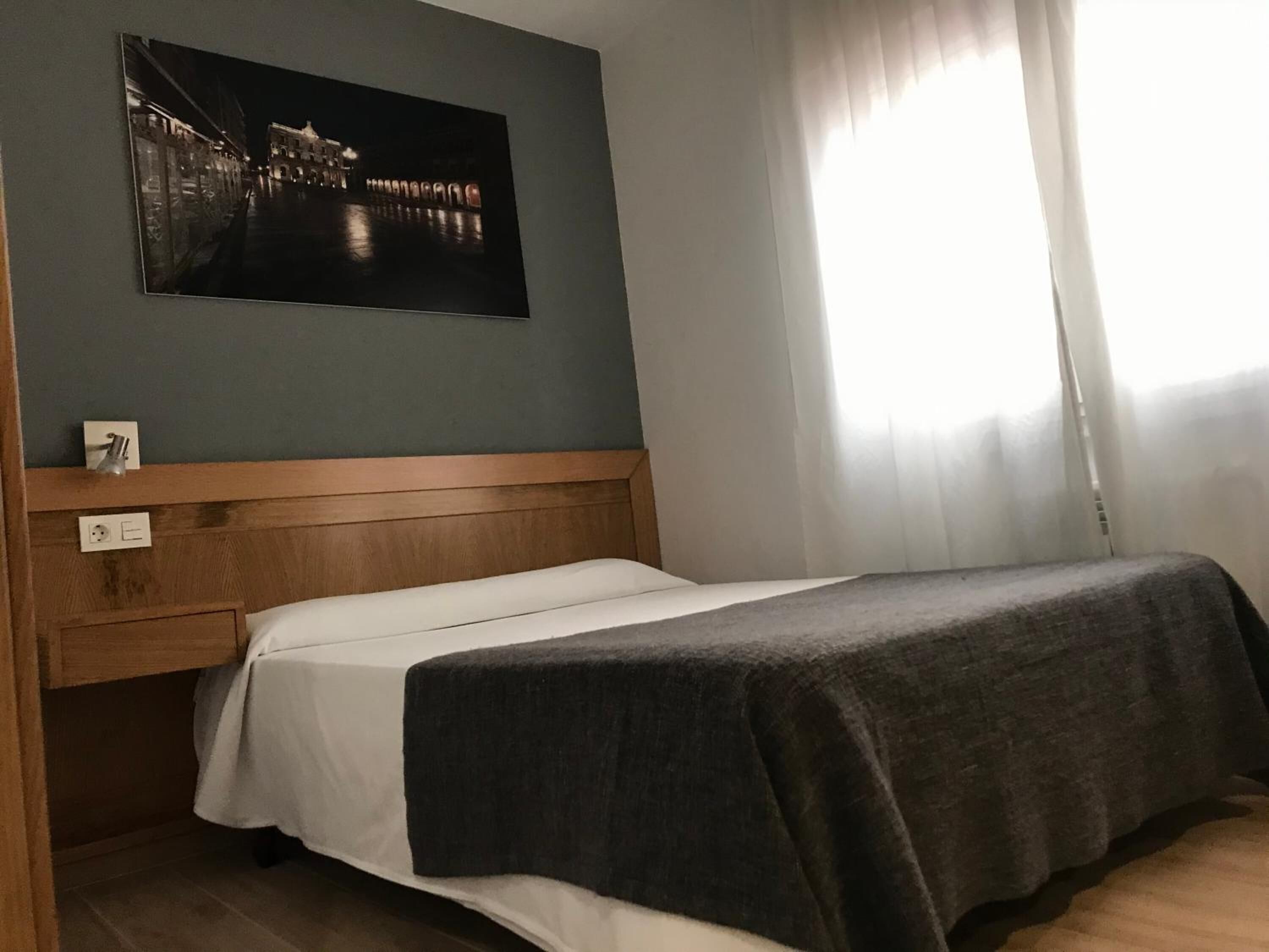 Une chambre avec un lit, une tête de lit en bois, un interrupteur fixé au mur et une fenêtre ornée de rideaux légers.