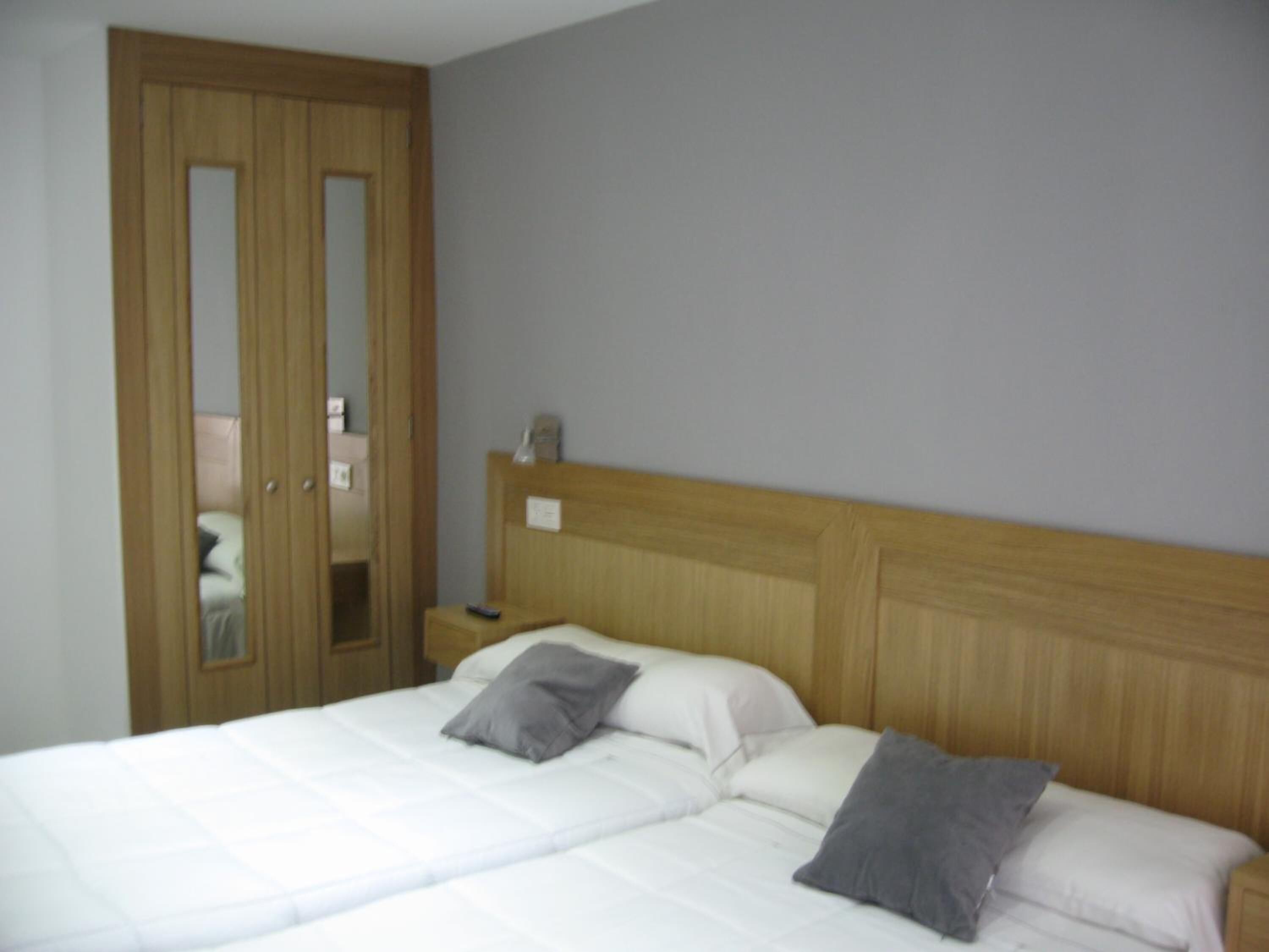 Quarto com cabeceira de madeira, cama com roupa de cama branca e duas almofadas, mesa de cabeceira com abajur e guarda-roupa de madeira com espelho.