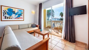 A modern hotel room with a sofa, a small table, a TV, and a balcony with a view of palm trees. - Vibra Blanc Palace Aparthotel (Ciutadella de Menorca)