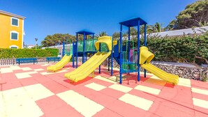 A playground with multiple slides and climbing structures, surrounded by a checkered pavement and a yellow building in the background. - Vibra Blanc Palace Aparthotel (Ciutadella de Menorca)