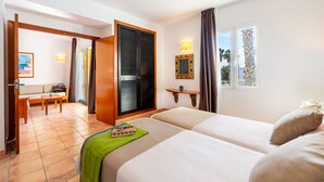 A hotel room with two beds, a sitting area with a sofa and table, a wooden door, a wall-mounted shelf, and a window with curtains. - Vibra Blanc Palace Aparthotel (Ciutadella de Menorca)