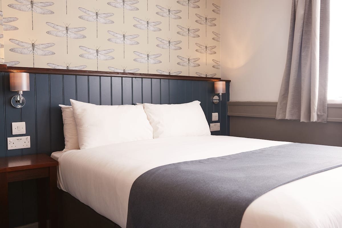 Une chambre d’hôtel comprenant un lit, deux tables de chevet, une tête de lit, des lampes fixées au mur et une fenêtre avec des rideaux.