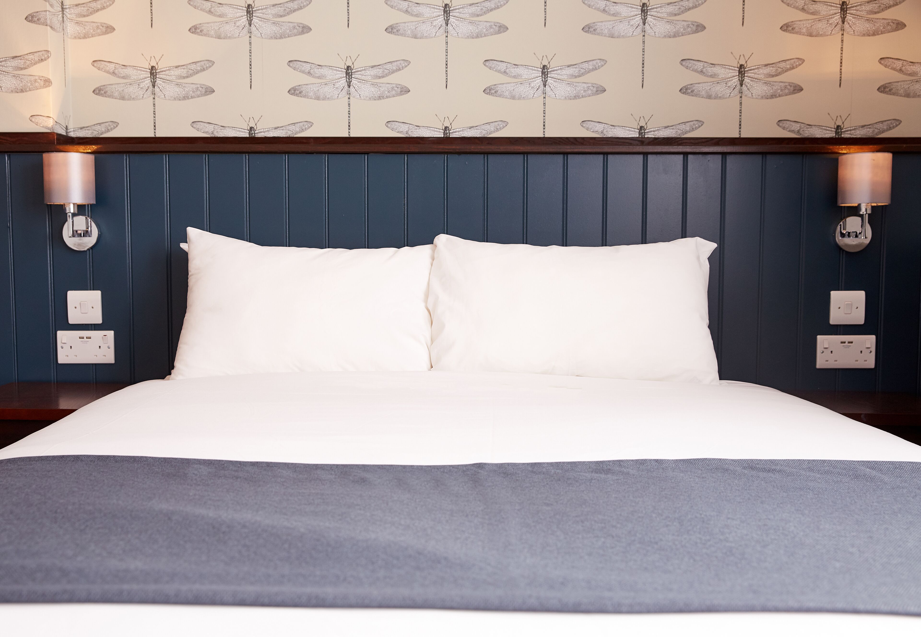 A neatly made bed with white linens and a gray throw blanket, a dark blue headboard with vertical paneling, and wall-mounted sconces with a dragonfly pattern wallpaper above.