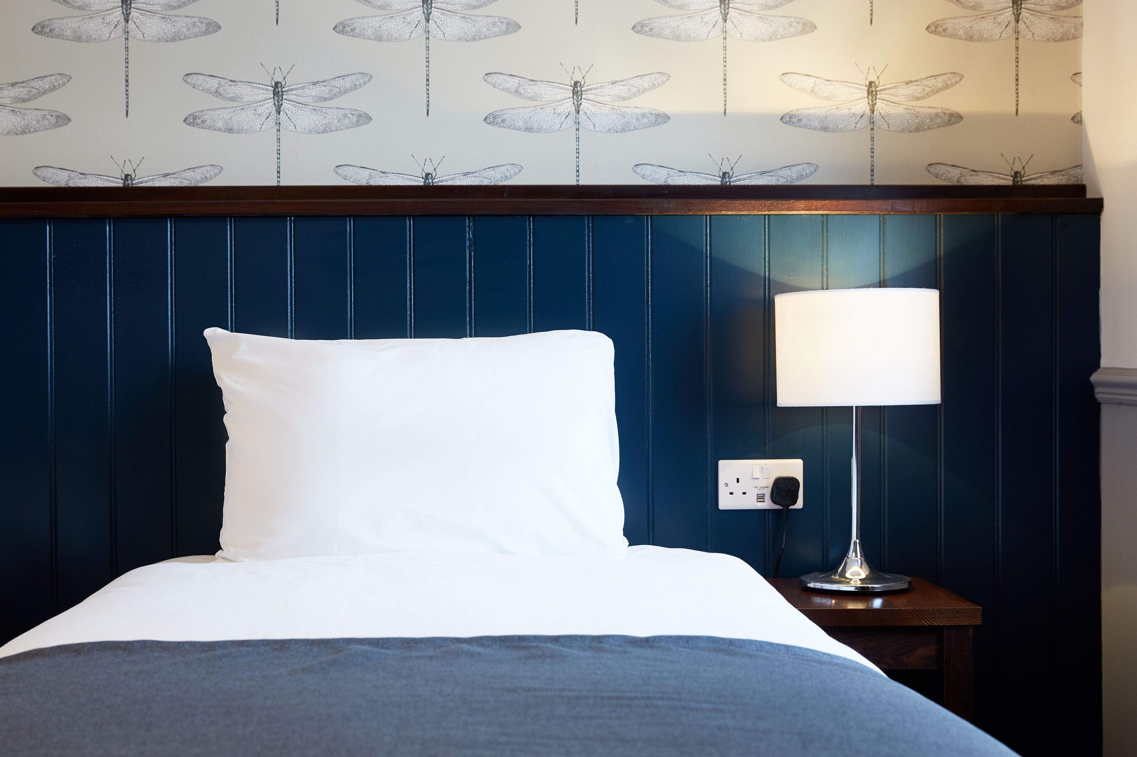 A neatly made bed with white bedding, a blue headboard, and a bedside table with a lamp.