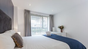 Une chambre d’hôtel moderne avec un grand lit, une table de chevet ornée d’un vase et une vue sur la ville à travers de fines rideaux.