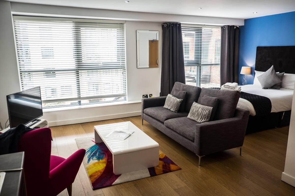 Une chambre d’hôtel moderne avec un canapé gris, une table basse blanche, un fauteuil rouge, un lit avec une tête de lit noire et de grandes fenêtres équipées de stores.