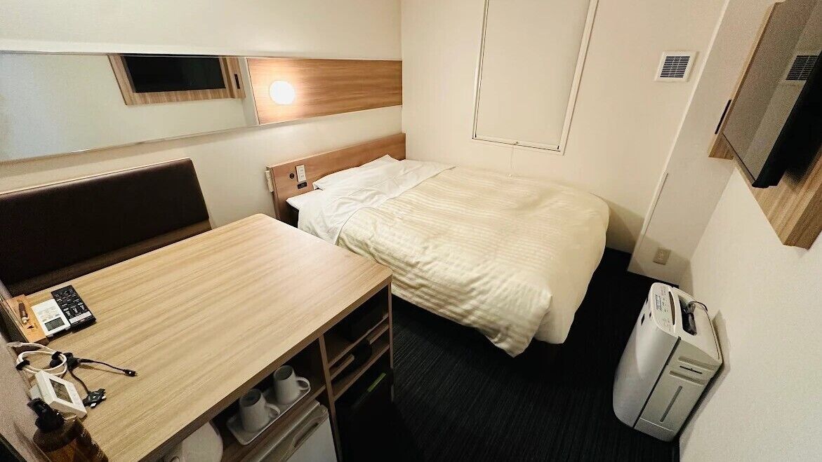 A compact hotel room with a bed, a small desk, a chair, a bedside table, a heater, and a television.