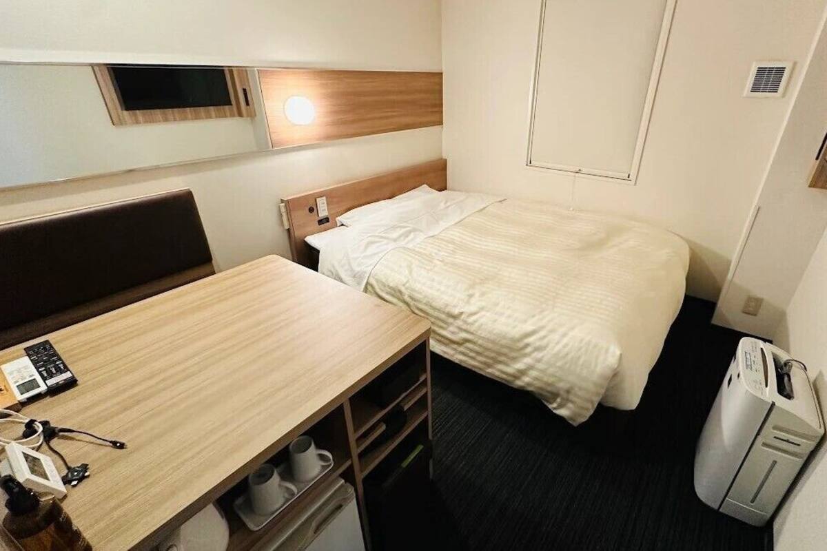 A compact hotel room with a bed, a small desk, a chair, a bedside table, a heater, and a television.
