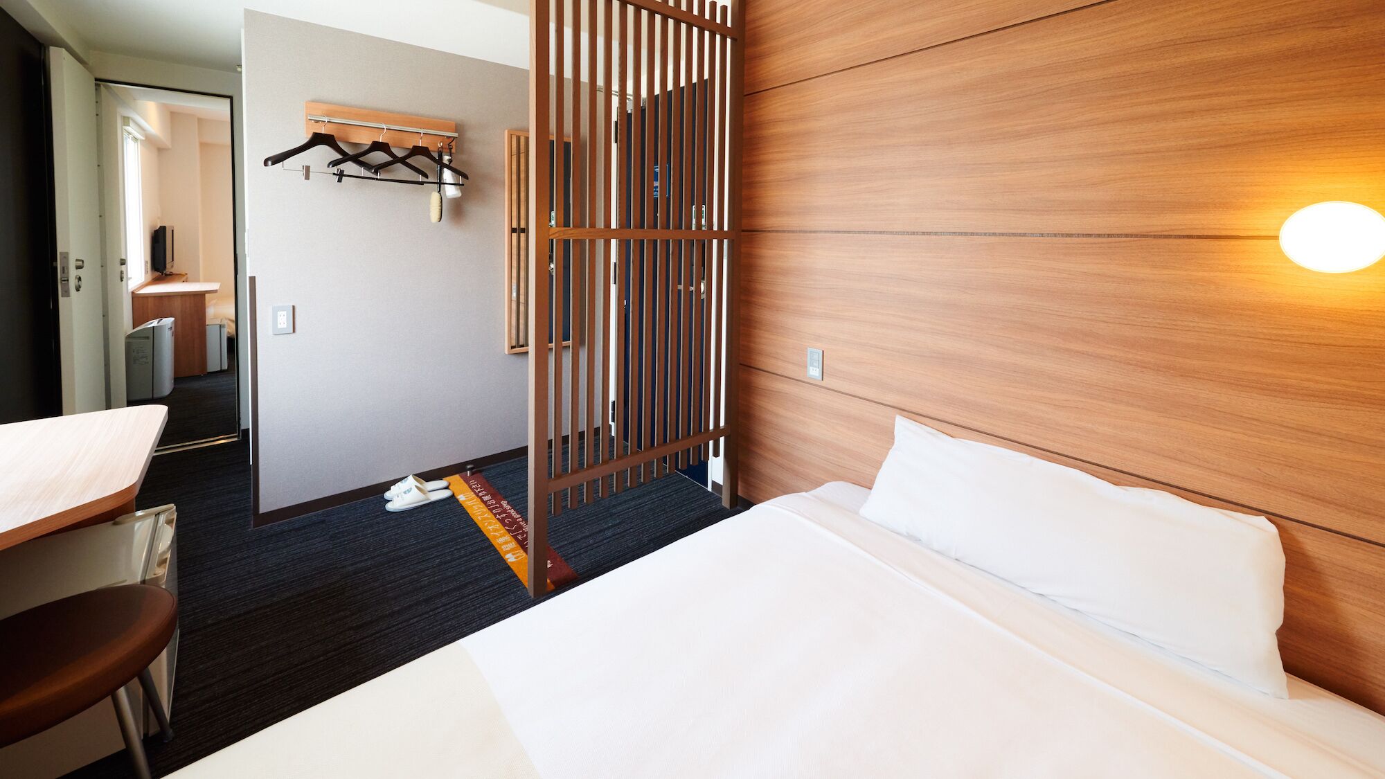 Habitació d'hotel amb llit, escriptori, cadira, armari i un separador amb llum.