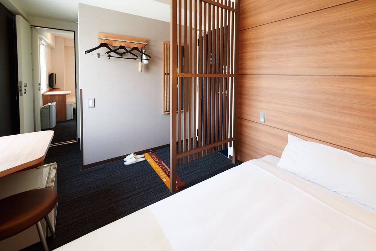 A hotel room with a bed, a desk, a chair, a wardrobe, and a partition with a light.