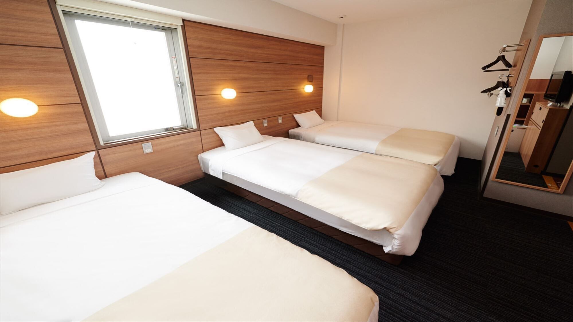 A hotel room with three beds, a large window, a mirror, and a wardrobe with hanging clothes.