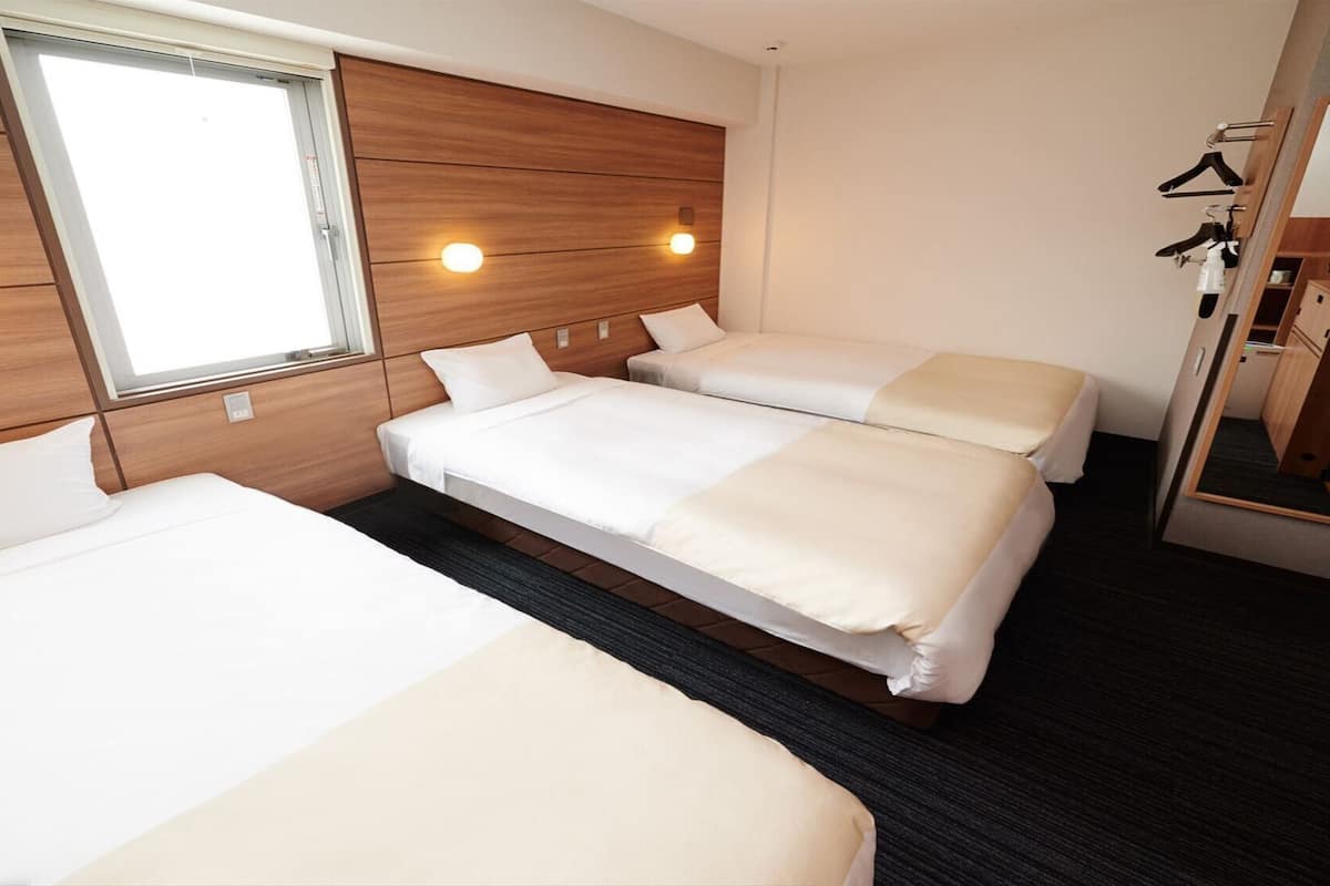 A hotel room with three beds, a large window, a mirror, and a wardrobe with hanging clothes.