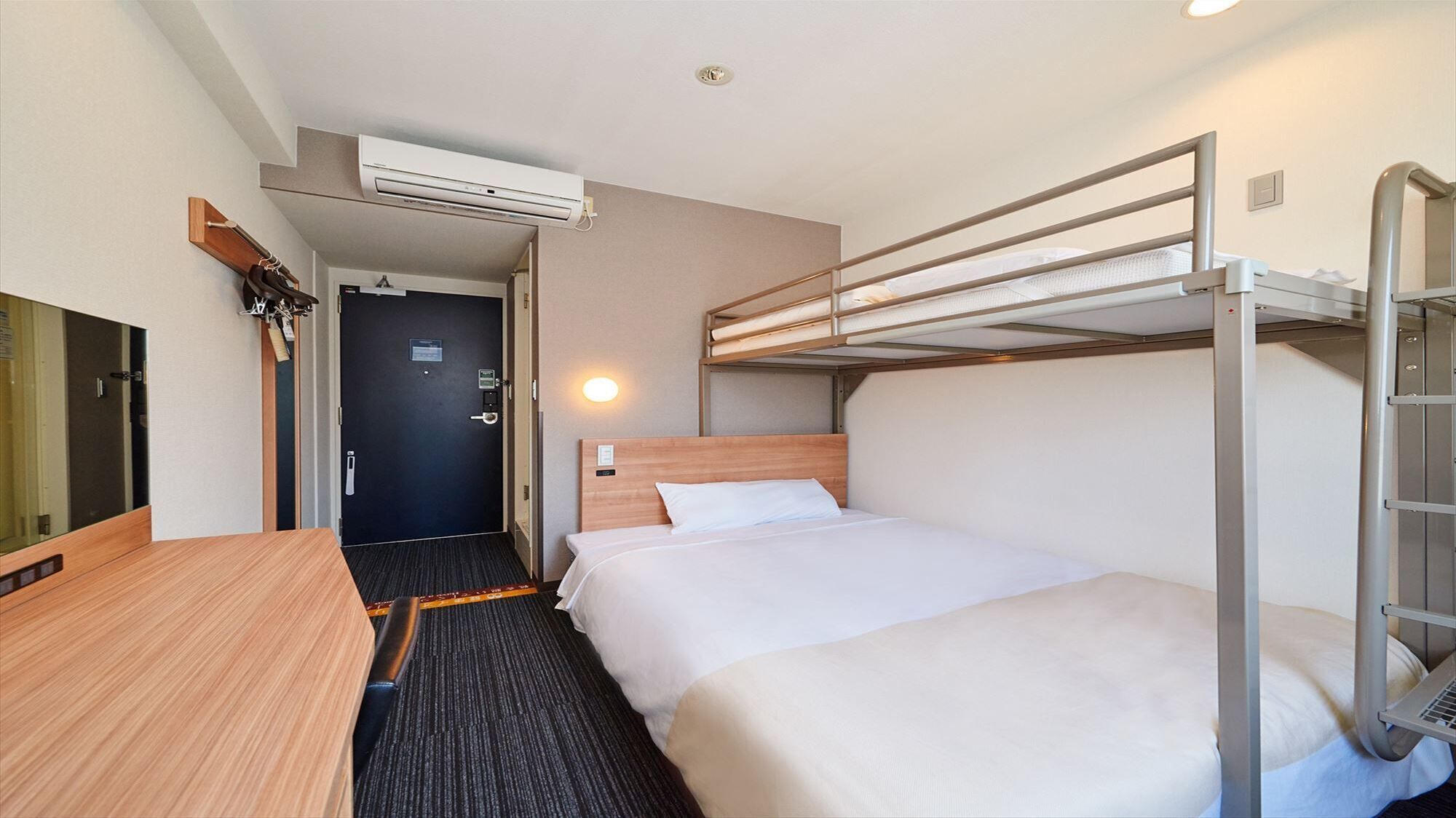 A hotel room with a bunk bed, a desk, a chair, a mirror, an air conditioning unit, and a door.