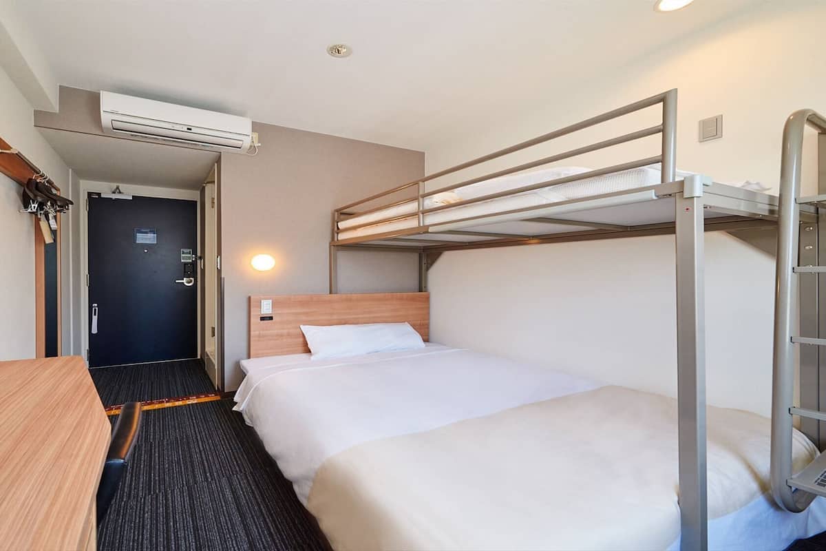 A hotel room with a bunk bed, a desk, a chair, a mirror, an air conditioning unit, and a door.