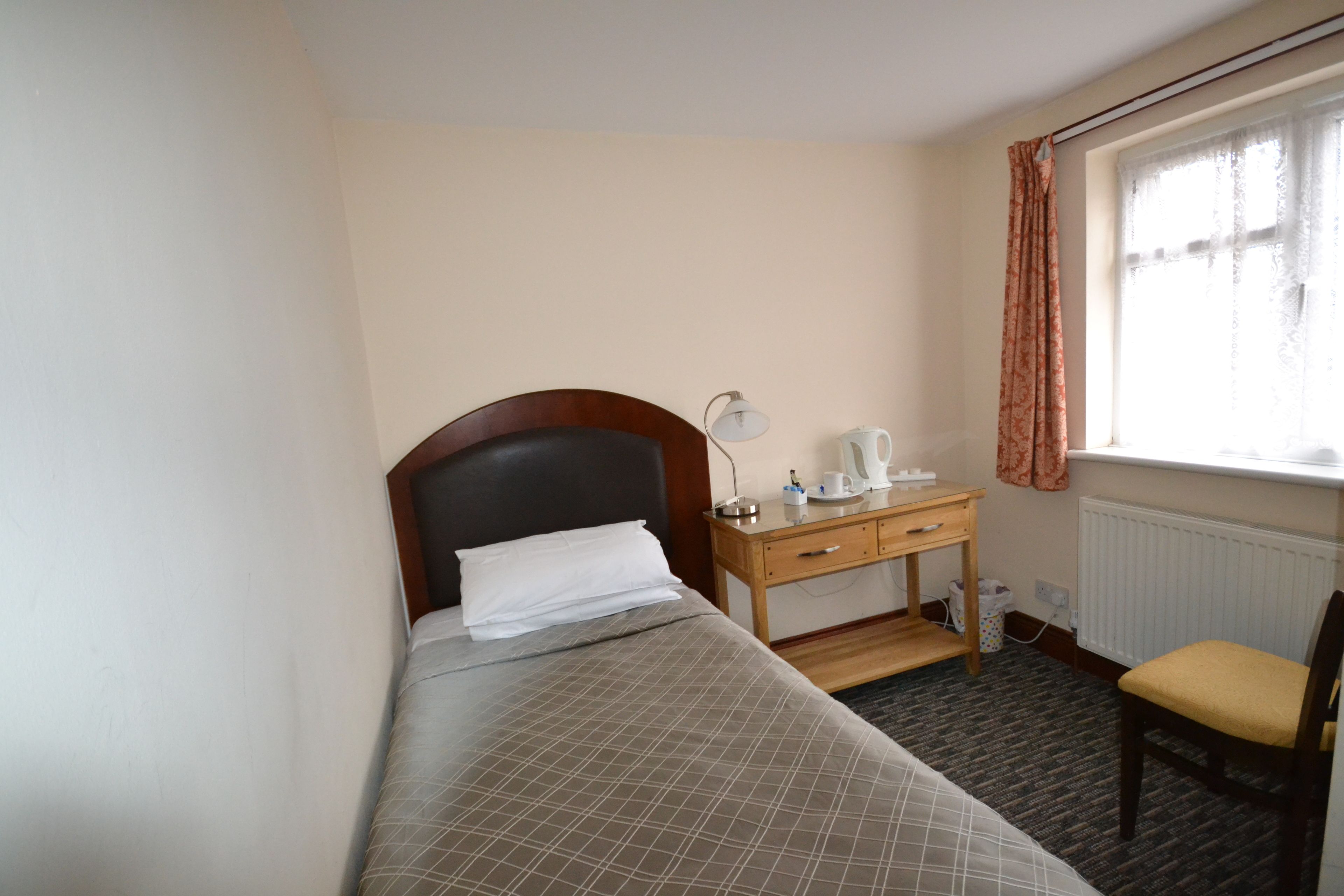 A small hotel room with a single bed, a wooden desk with a lamp, a chair, and a window with curtains.