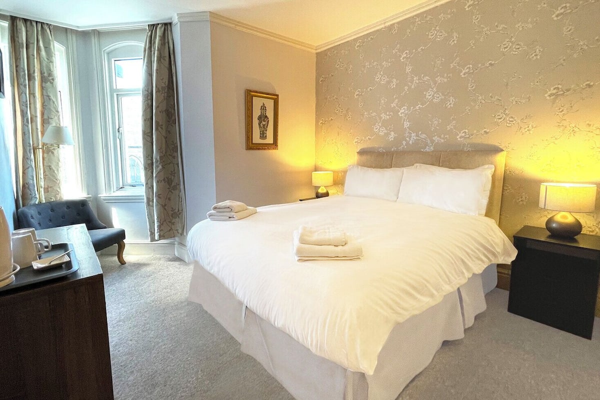 A hotel room with a large bed, two bedside tables with lamps, a chair, a desk with a telephone, and a window with curtains.