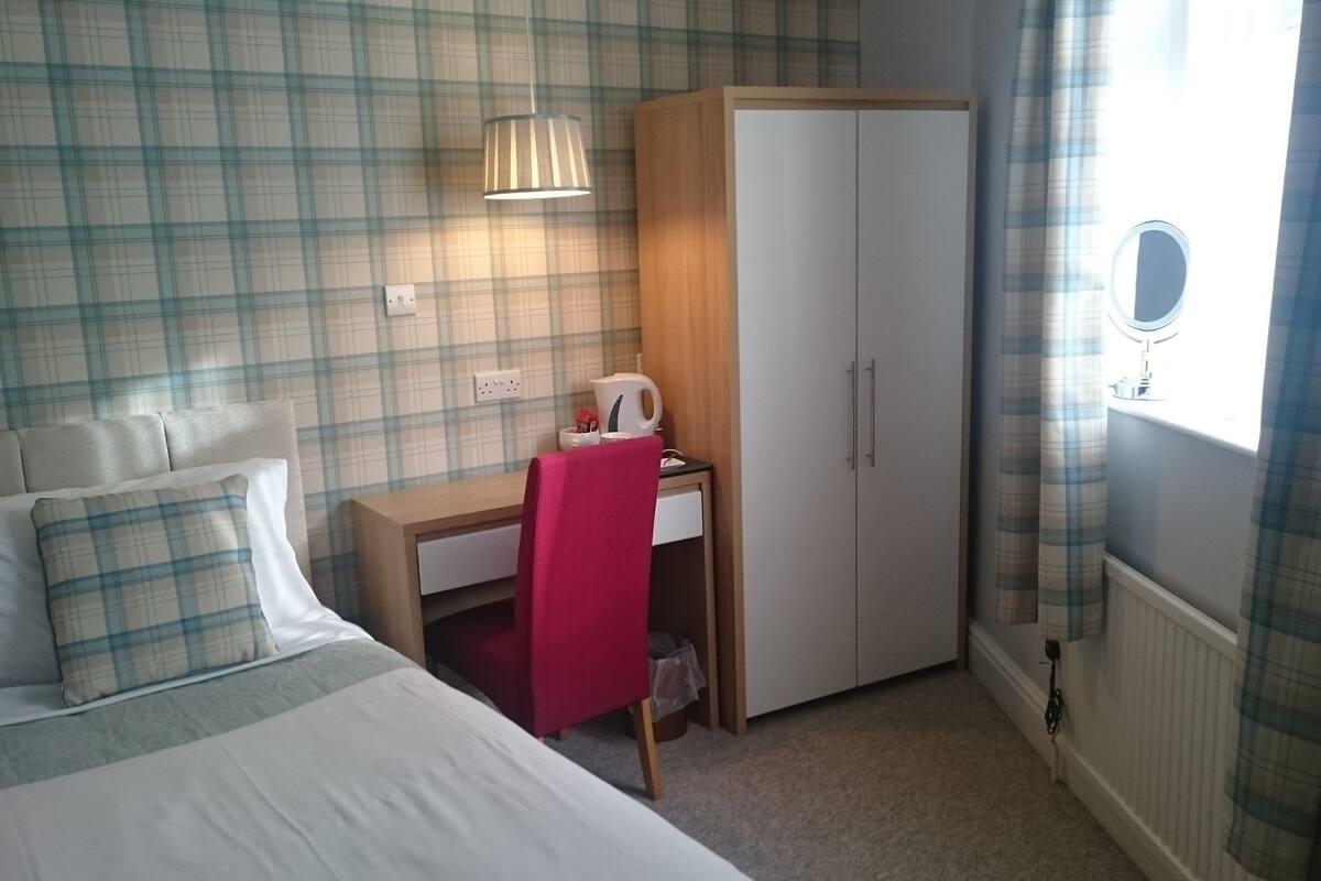 A bedroom with a bed, a desk, a wardrobe, a window with curtains, and a wall-mounted lamp.