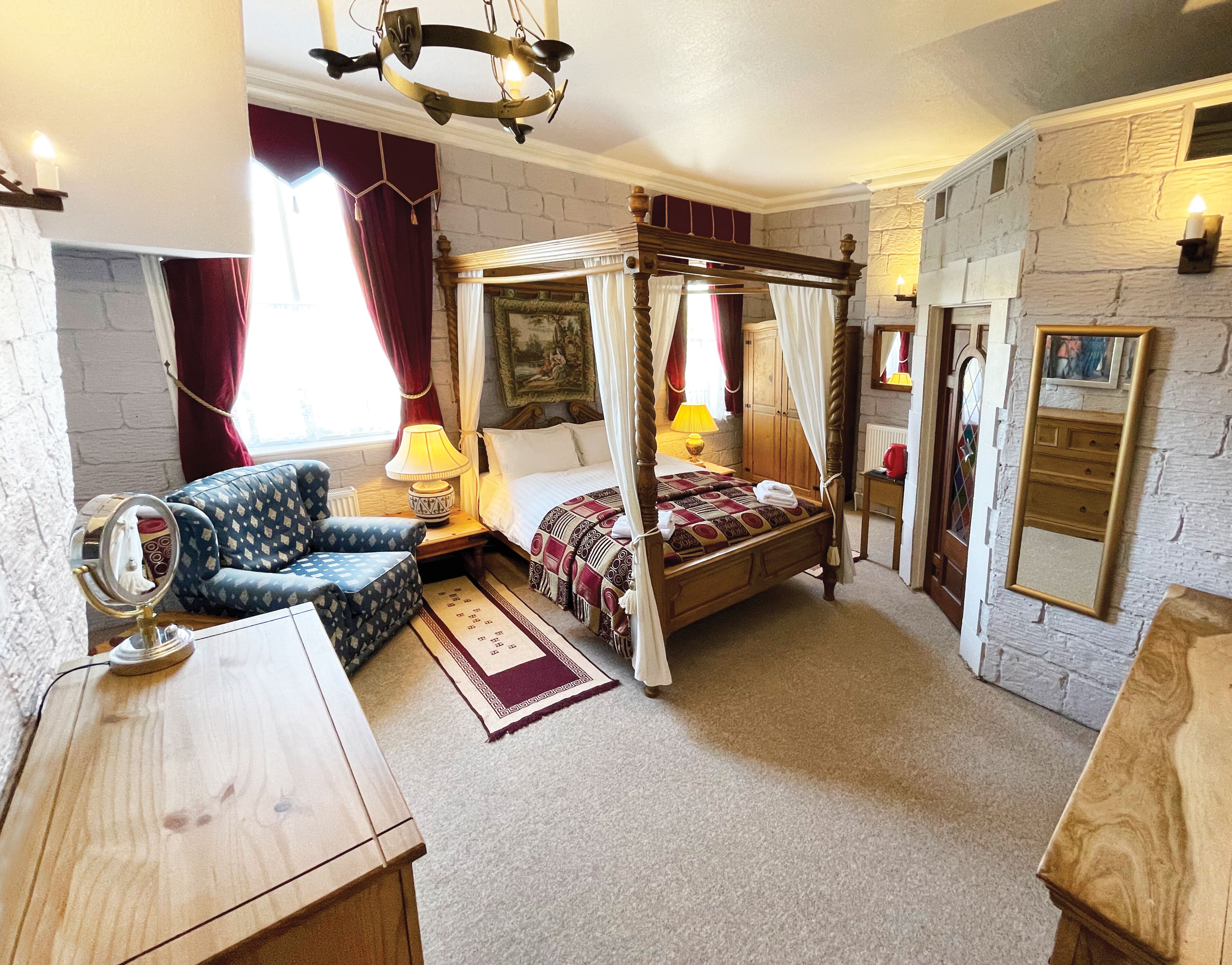 A bedroom with a four-poster bed, a wooden nightstand, a chair, a mirror, and a stone wall.