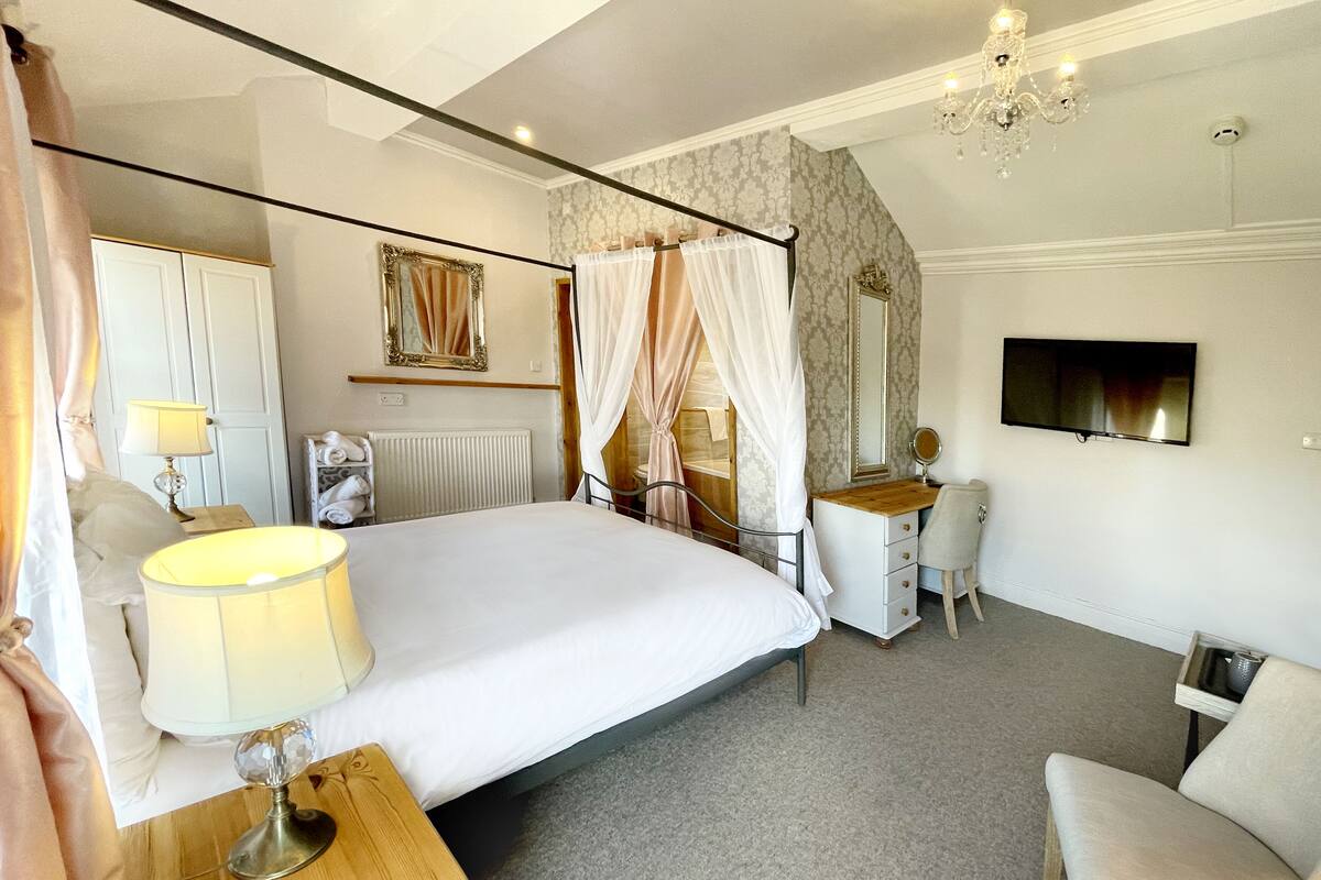 A bedroom with a large bed, a bedside table with a lamp, a vanity with a mirror, a chair, and a television on the wall.