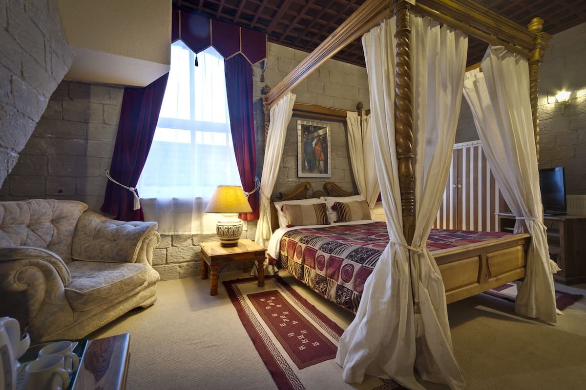 A bedroom with a four-poster bed, a wooden nightstand, a lamp, a sofa, and a stone wall.