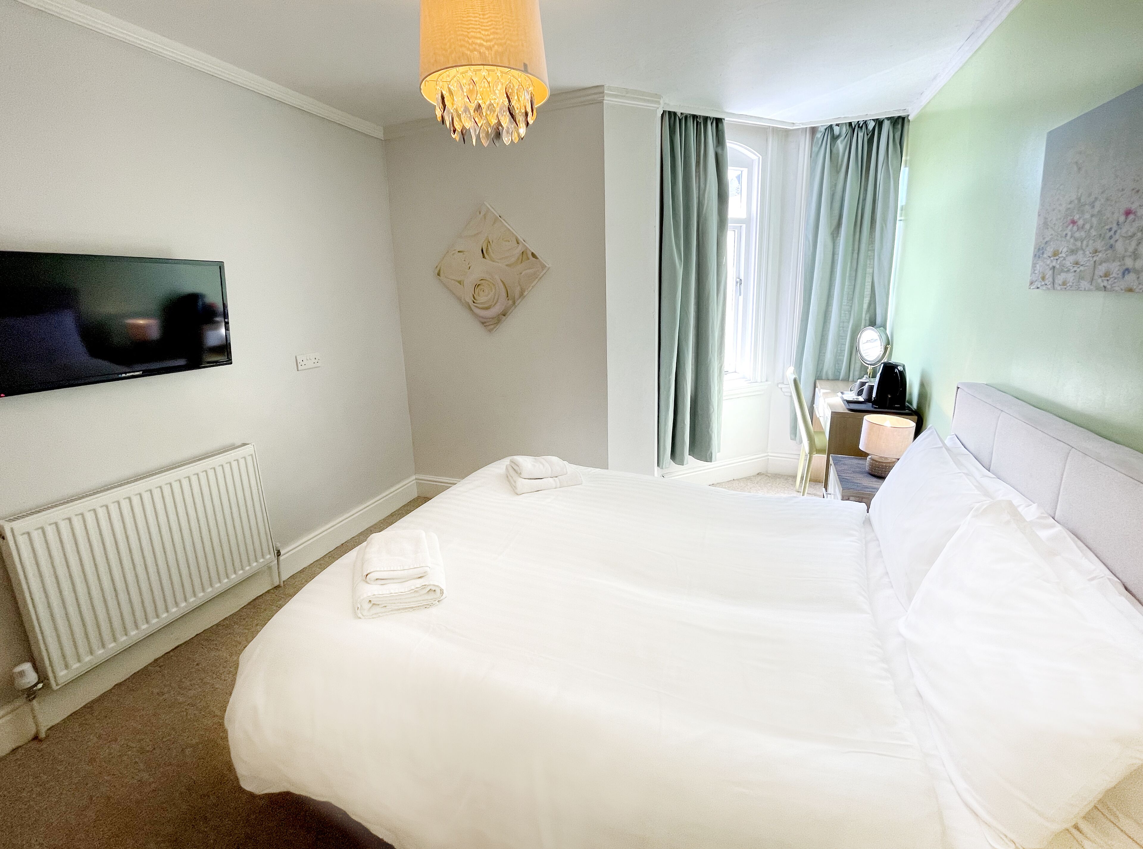 Ein Hotelzimmer mit einem großen Bett, einem an der Wand montierten Fernseher, einem Heizkörper, einem Schreibtisch mit Lampe sowie einem Fenster mit Vorhängen.